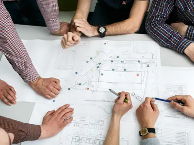 Table With Blueprints Hands Holding Pencils